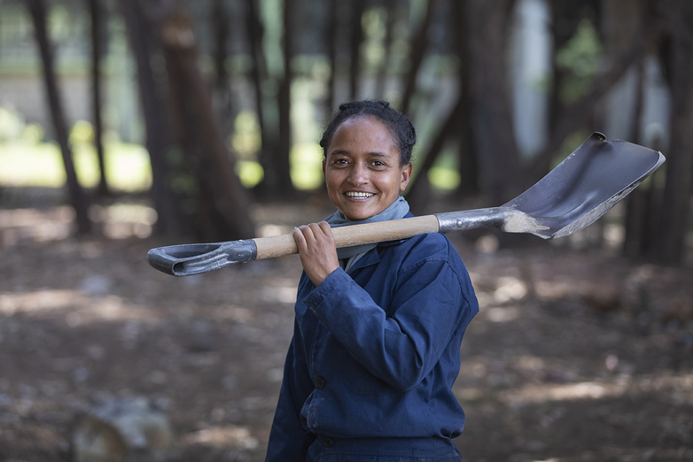 A woman carries a spade over her shoulder and smiles into the camera.