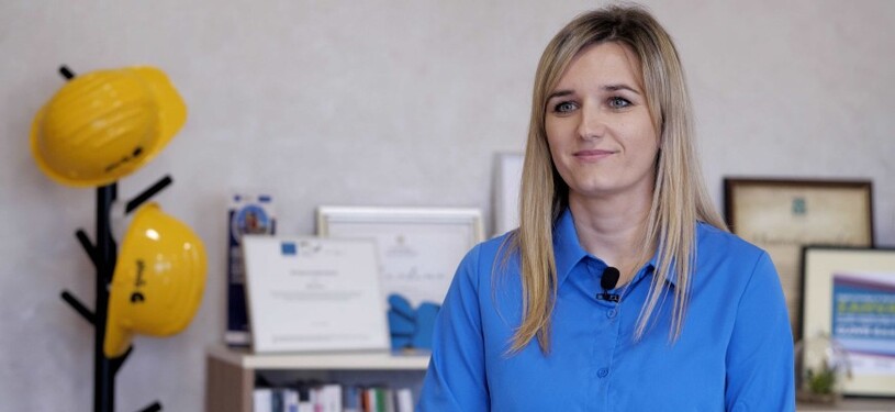 Naida Baručija sits in front of a shelf on which a number of framed certificates are displayed.