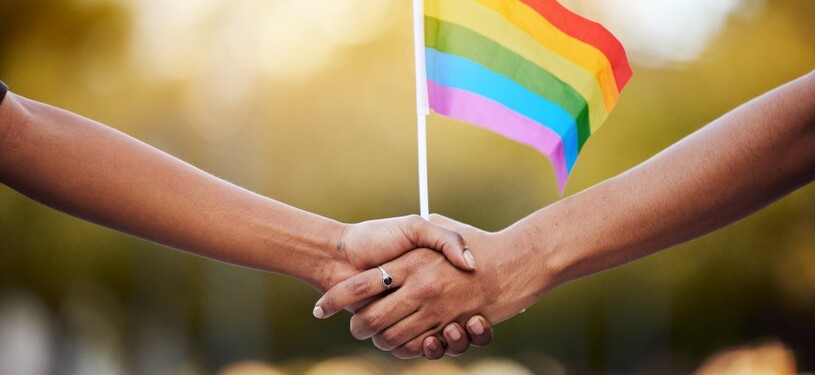 Two people shake hands while holding a rainbow flag together.