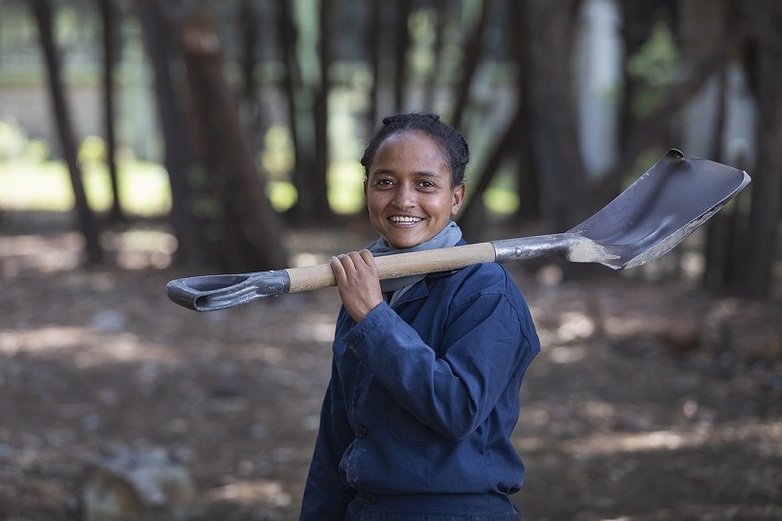 A woman carries a spade over her shoulder and smiles at the camera. Copyright: GIZ
