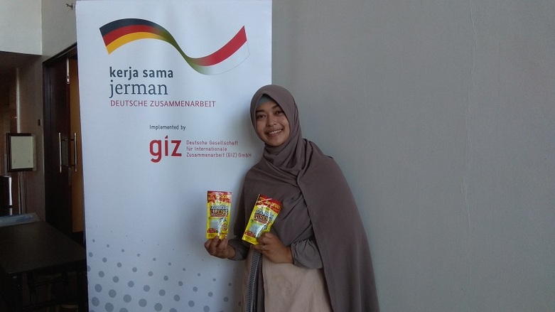 A woman wearing a hijab is holding two packages from the diaspora start-up 'Mak Rah Pireng' in her hands in front of a GIZ stand-up display. Copyright: GIZ