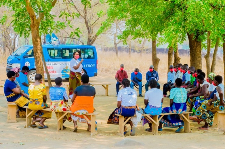 Several people wearing protective masks sit in a circle and listen to a woman. In the background is a minibus with satellite dishes on the roof. Copyright: GIZ