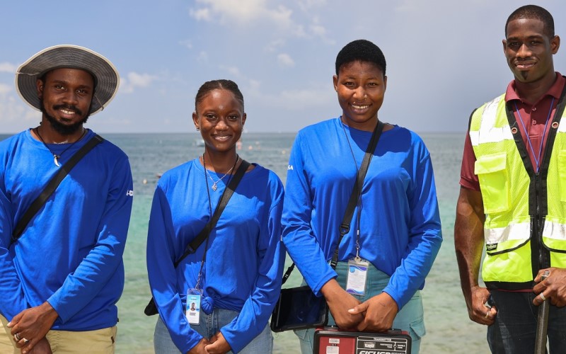 Four people in working gear standing on the beach with the ocean in the background look directly into the camera.