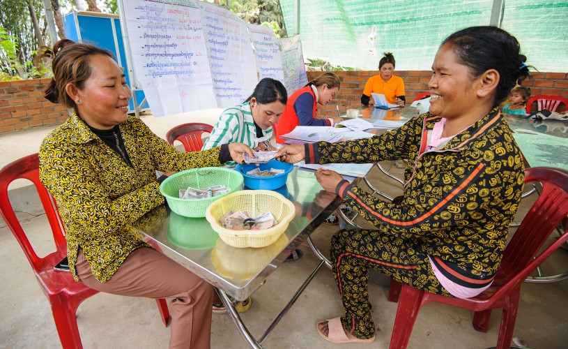 A woman hands banknotes to another woman sitting opposite her at a table. There are bowls containing other banknotes on the table between them. In the background, three more women are writing in notebooks.