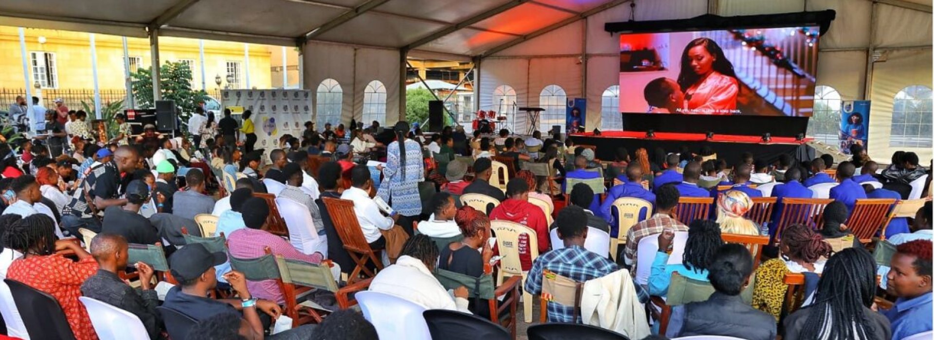 Around 150 people sit in a big tent watching a screen showing a scene from a film.